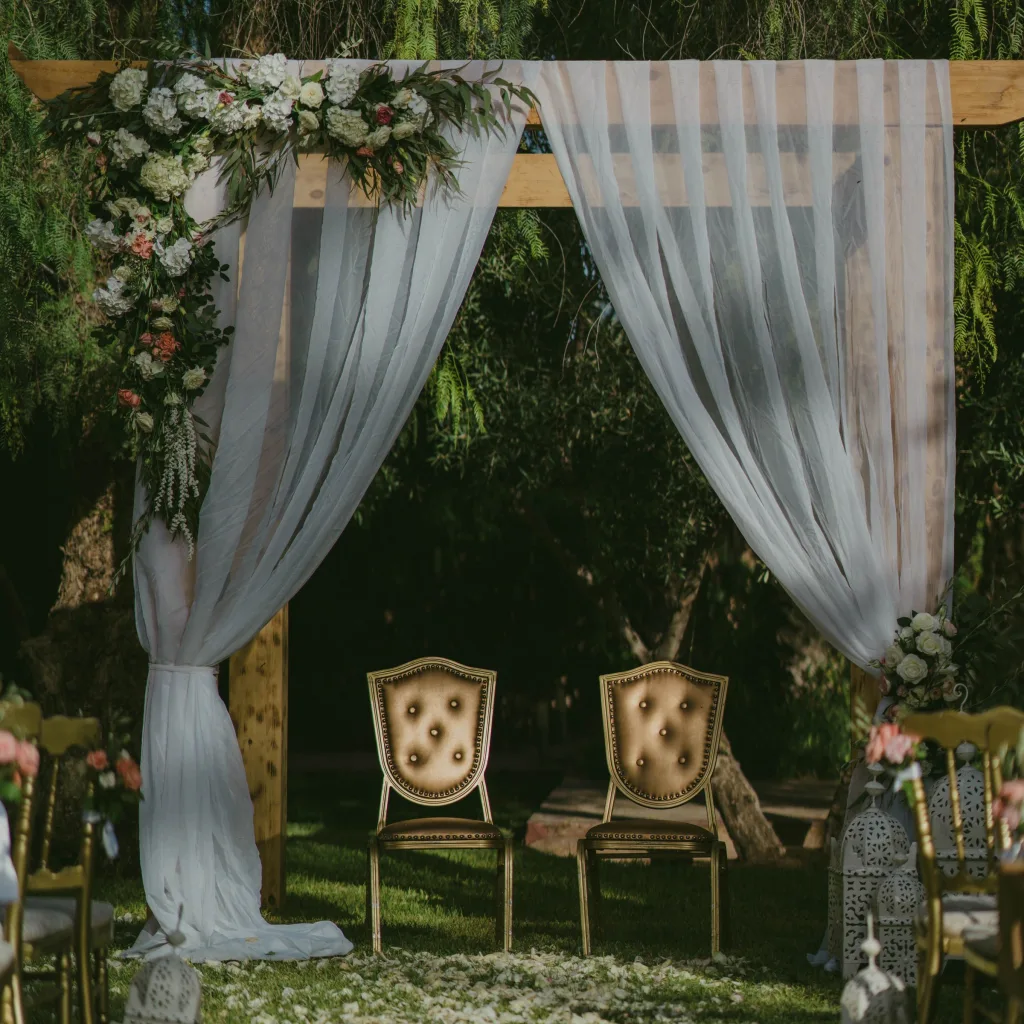 Arche de cérémonie drapée de voilages et de fleurs blanches, deux fauteuils dorés pour les mariés, dans un jardin à Marrakech