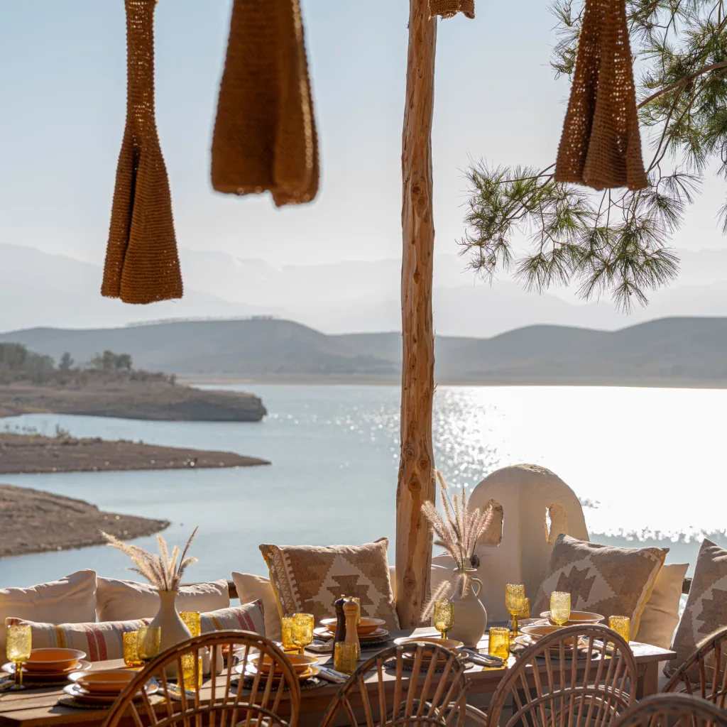 Longue table dressée avec banquettes et coussins, au bord d’un lac, pour un repas de groupe
