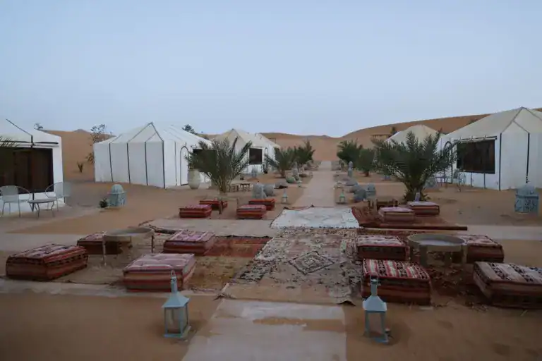 Salon en plein air d’un camp dans les dunes : tapis, poufs et lanternes entre les tentes blanches
