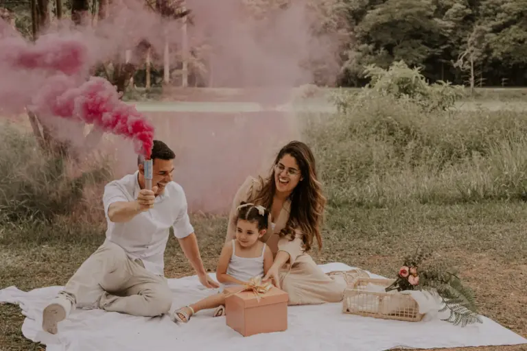 Couple et sa fillette assis sur une nappe en plein air, fumigène rose et paquet cadeau pour une révélation du sexe du bébé