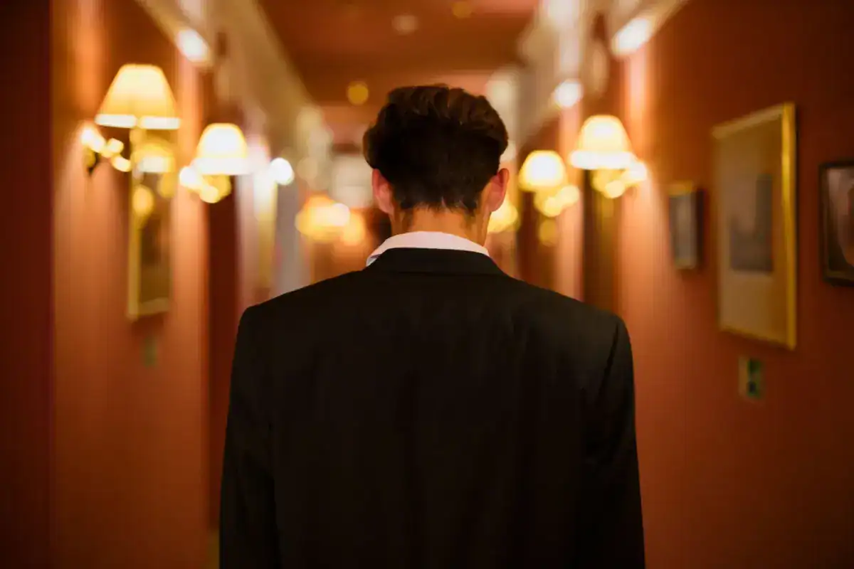 Homme en costume sombre, de dos, dans un couloir d’hôtel éclairé par des appliques