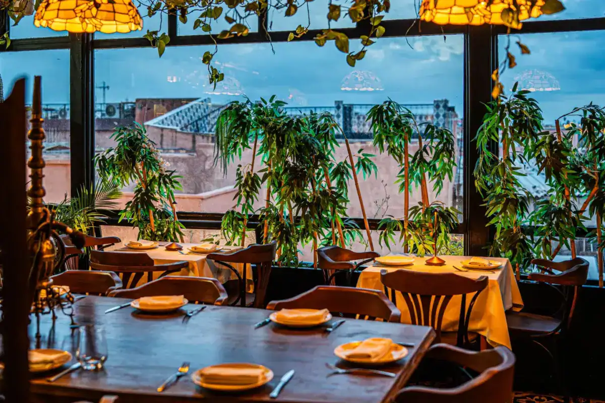 Salle de restaurant aux tables dressées sous des suspensions ambrées et des plantes, avec vue sur les toits de la ville