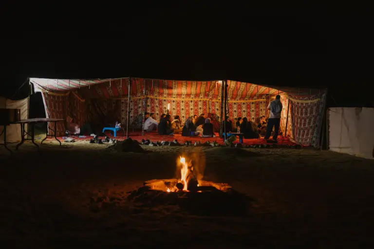 Invités réunis sous une tente de bivouac éclairée, feu de camp dans le sable à la nuit tombée