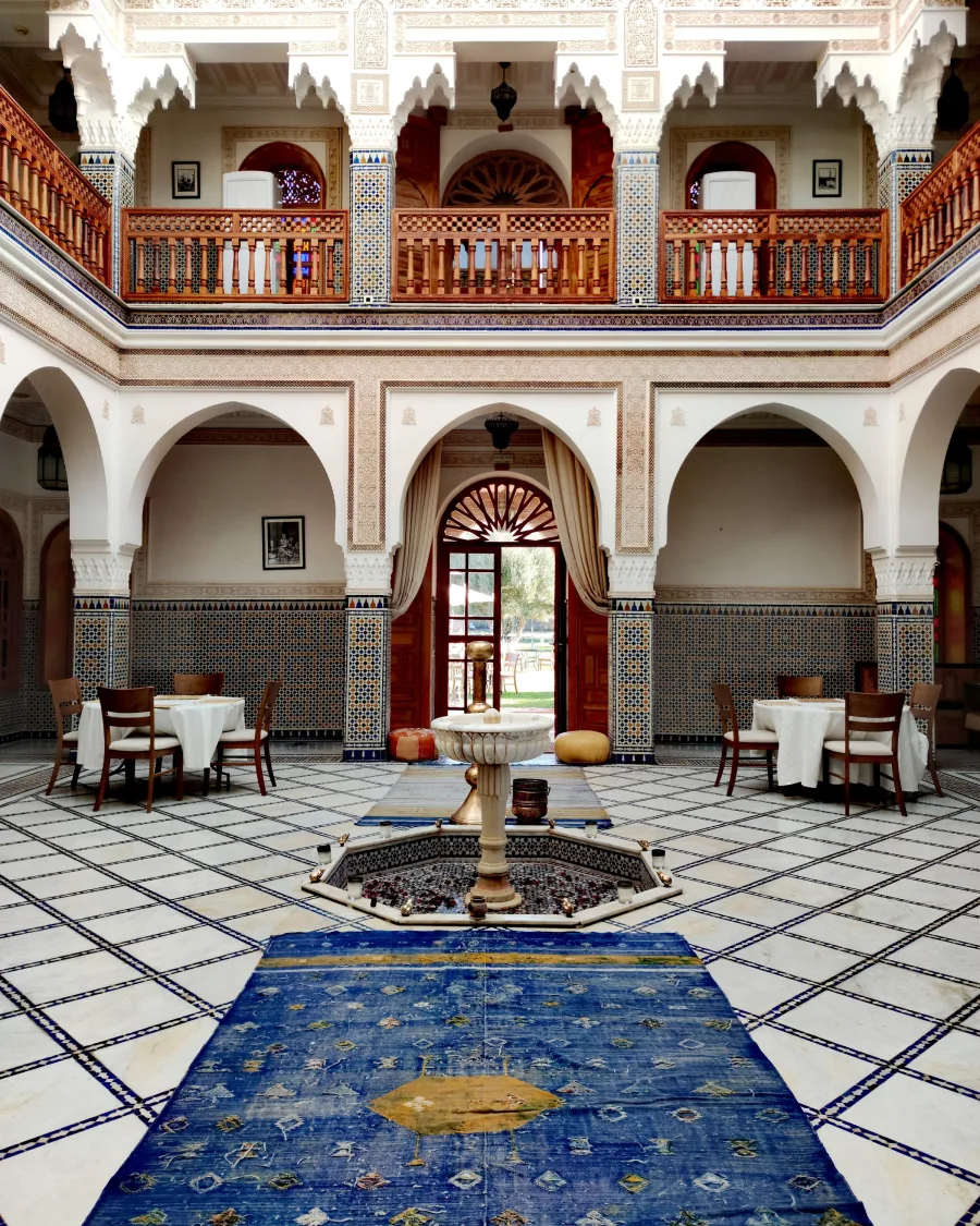 Courtyard of a grand Moroccan house set for dinner: white-clothed tables, zellige tiles and a central fountain