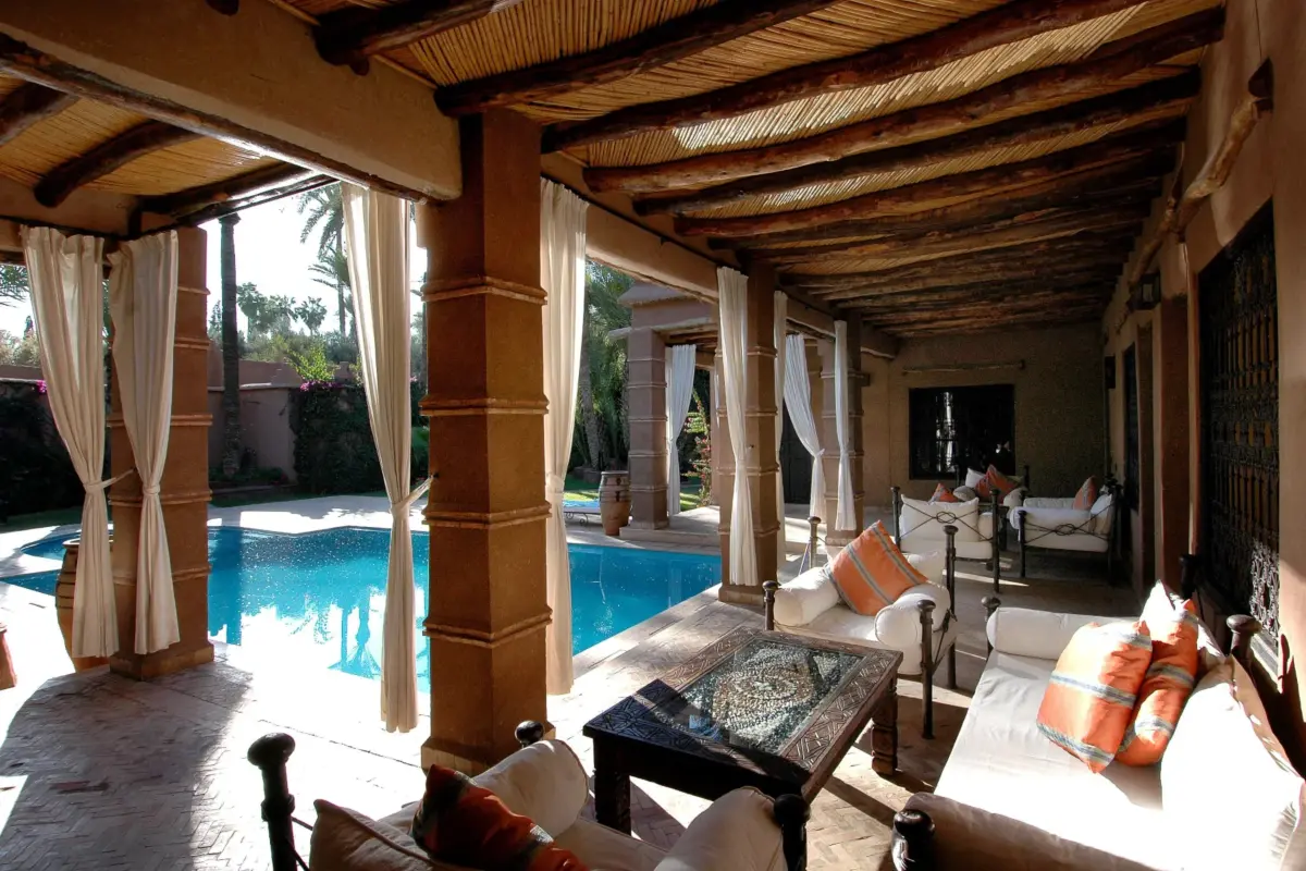 Terrasse couverte d'une villa de style marocain avec salon, rideaux et piscine dans un jardin de palmiers