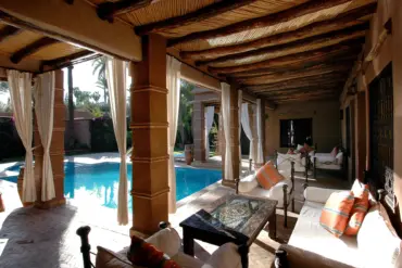 Terrasse couverte d'une villa de style marocain avec salon, rideaux et piscine dans un jardin de palmiers