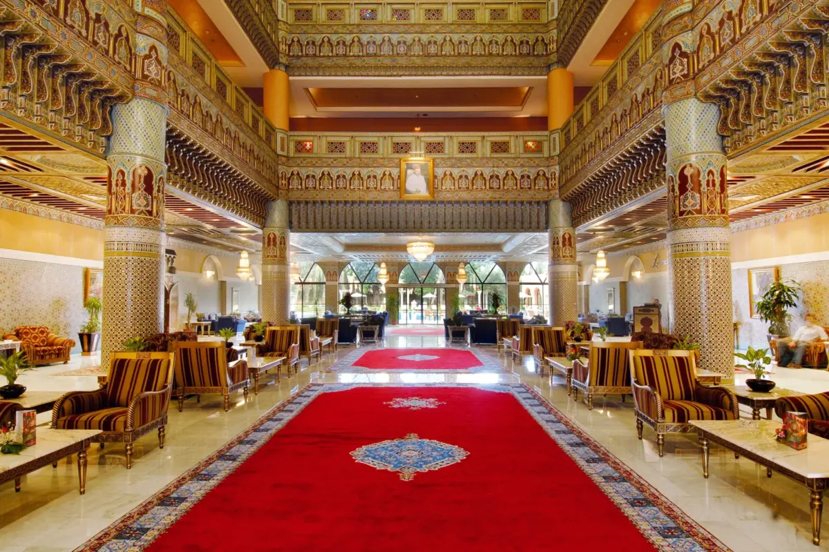 Grand salon d'un hôtel marocain avec tapis rouge, colonnes en zellige et plafonds en bois sculpté