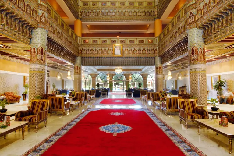 Grand salon d'un hôtel marocain avec tapis rouge, colonnes en zellige et plafonds en bois sculpté