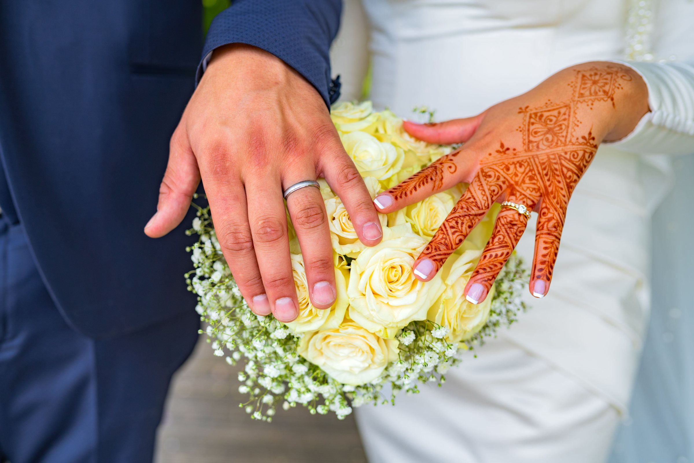 Mains d'un couple de mariés avec alliances et henné sur un bouquet de roses blanches