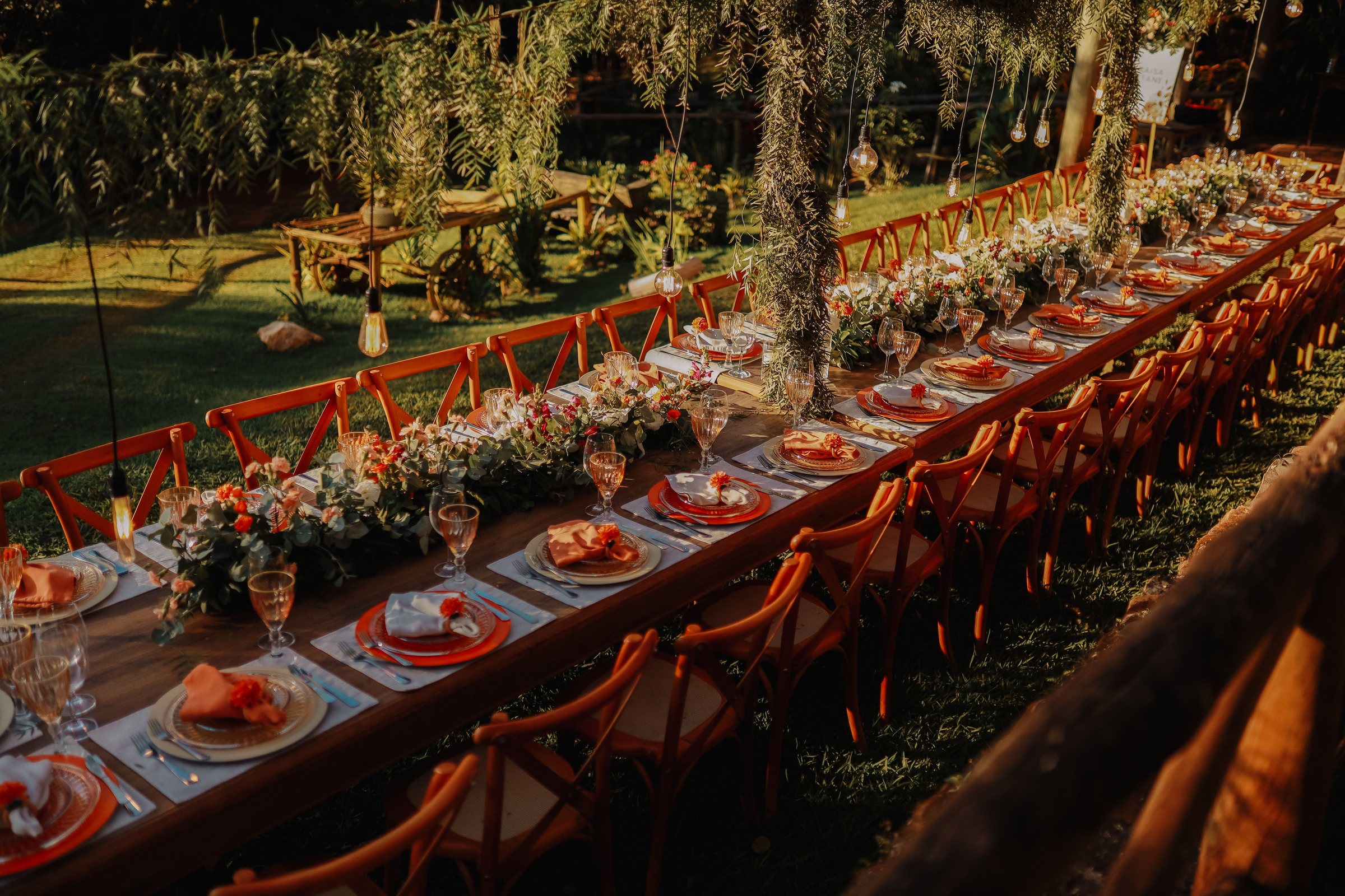 Longue table de dîner dressée dans un jardin, serviettes orange et fleurs