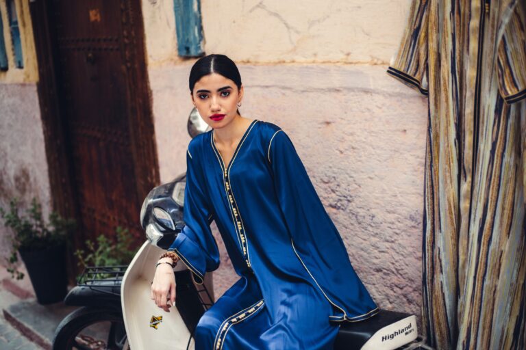 Femme en caftan bleu brodé assise devant une porte de médina