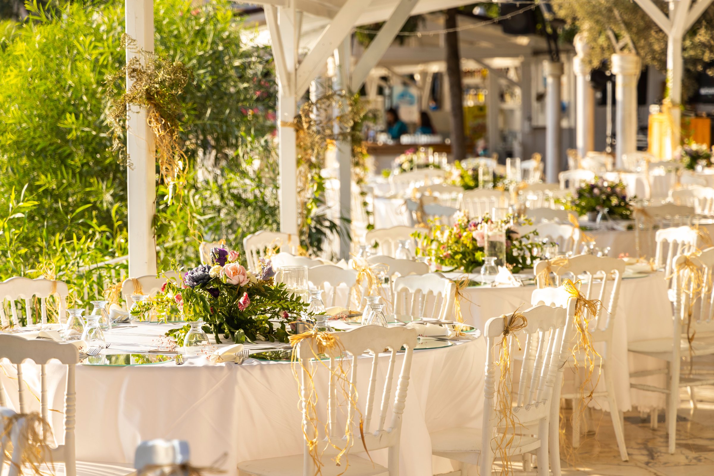 Réception en plein air, tables rondes, chaises blanches et fleurs sous une pergola
